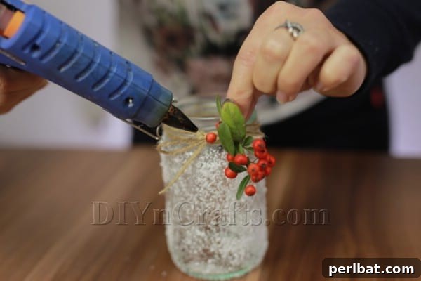 A small artificial holly sprig being hot glued onto the twine at the top of the frosted jar.