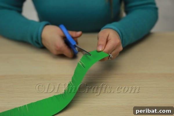 Green fabric or paper strip being cut into fringes with scissors.