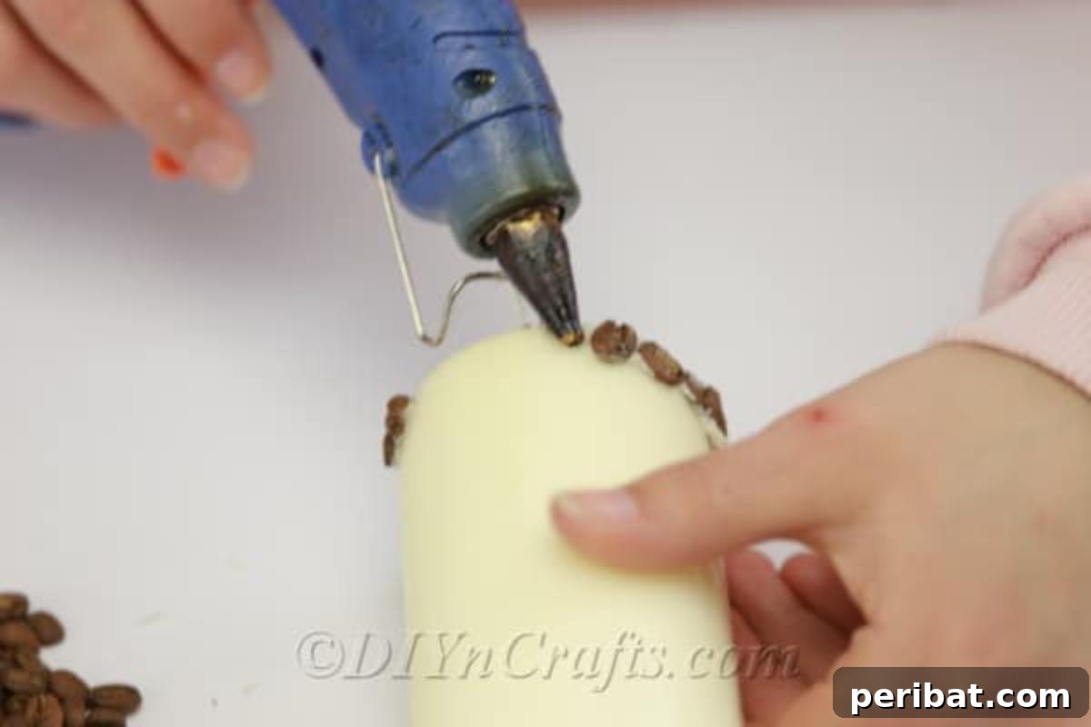 Demonstration of using a hot glue gun to attach individual coffee beans to a pillar candle, illustrating the precise application technique.
