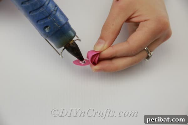 A hot glue gun is used to secure the central tab of a tightly rolled paper swirl, forming the base of a paper flower.