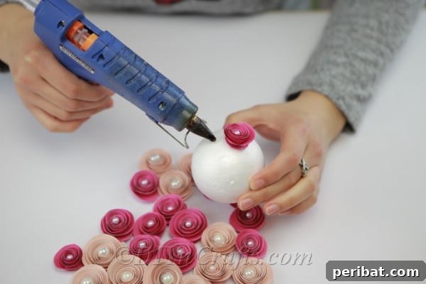 A hand meticulously gluing multiple finished paper flowers onto a Styrofoam ball using a hot glue gun, gradually covering the surface.