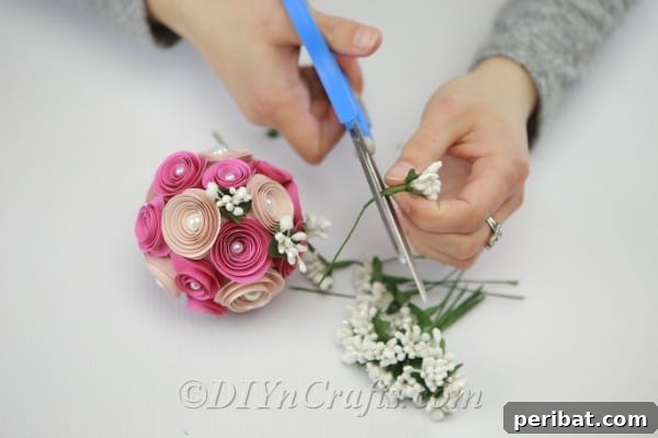 Delicate artificial branches and leaves being carefully glued into the gaps between paper flowers on a DIY floral ball, adding a natural touch.