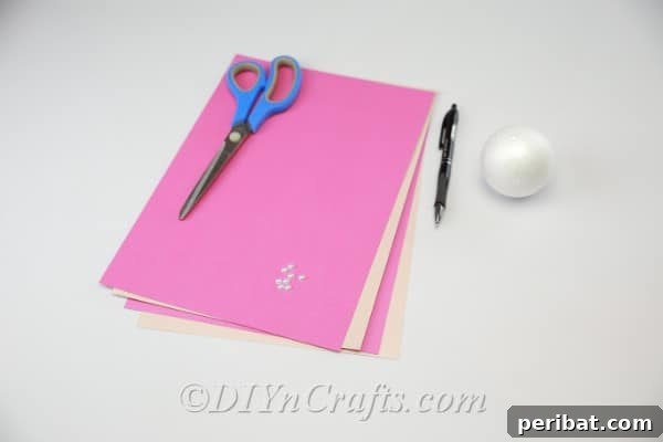 Various colored cardstock papers, a Styrofoam ball, pen, scissors, and a hot glue gun laid out as essential supplies for crafting a DIY paper flower ball.