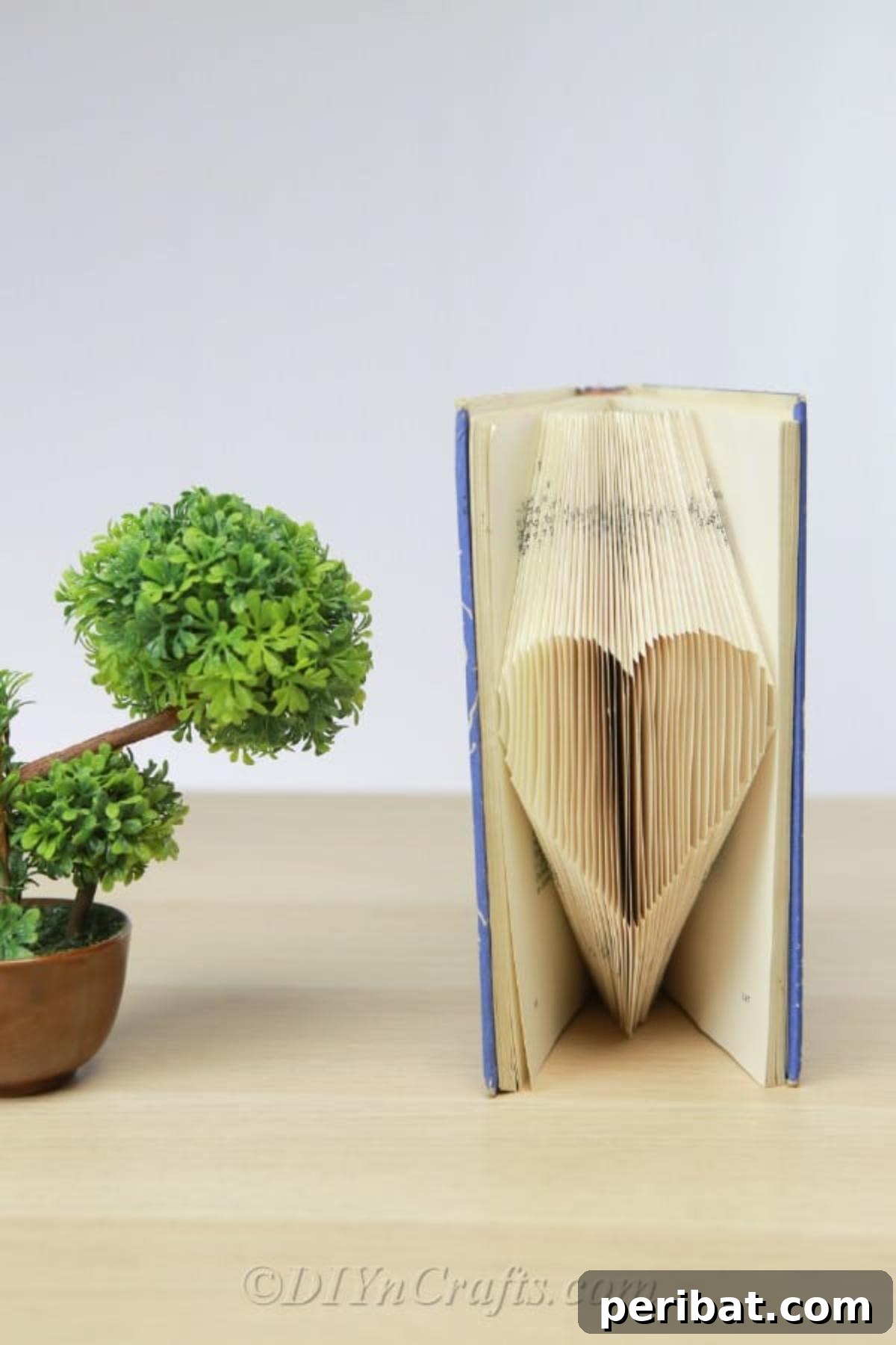A finished book art piece, showing a heart folded into the pages, displayed next to a small potted plant.