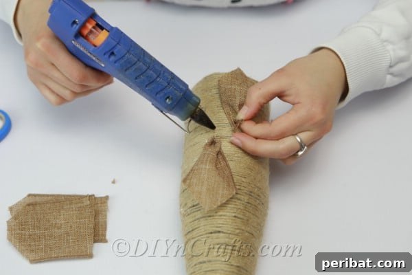Arranging and gluing burlap ribbon petals in a circular pattern onto the rope-covered bottle, forming a flower.