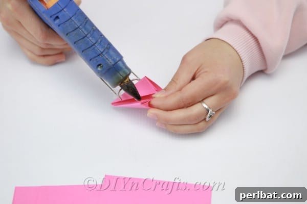 Applying hot glue to the overlapping edges of a pink paper petal to secure its folded shape
