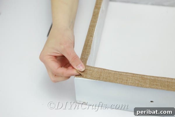 A person applying hot glue to secure a piece of burlap fabric onto the front of a rustic wooden box, adding a decorative touch to the DIY vase hanger.
