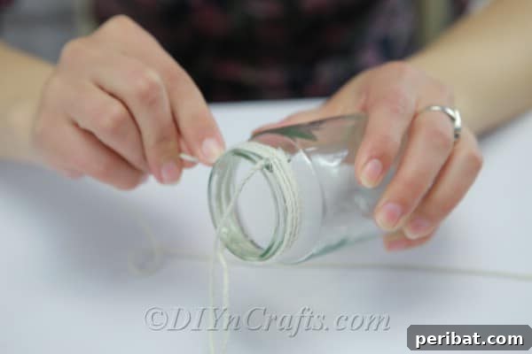 A close-up shot of a hand meticulously hot gluing natural twine around the neck of a glass jar, creating a loop for hanging in a DIY vase project.