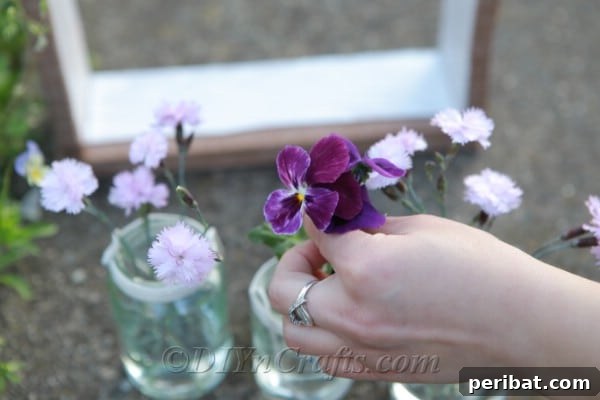 A hand gently placing fresh pink flowers into a glass jar suspended within a wooden box frame, illustrating the final step of arranging the DIY vase hanger.