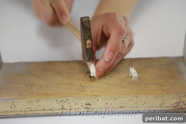A hand using a screwdriver to attach small hooks to the inside top edge of a rustic wooden box, ensuring they are securely fastened for hanging jars.