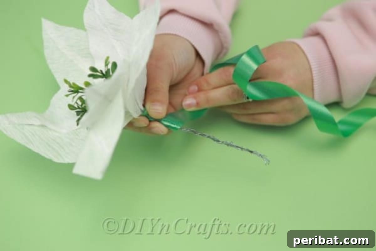A crafter meticulously wrapping green ribbon around the exposed floral wire at the base of a tissue paper flower, creating a natural-looking stem.