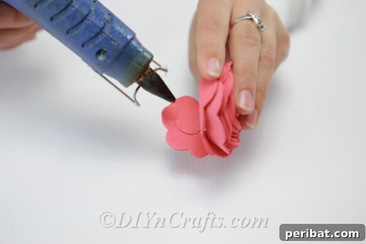 A crafter applies hot glue to the base of a partially rolled craft foam flower to secure its shape.