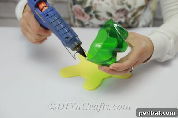 Applying hot glue to the rim of the plastic soda bottle bottom, then attaching it to the craft foam turtle body.