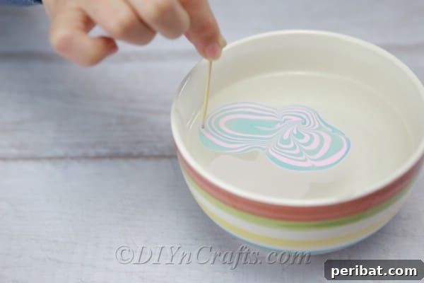 A toothpick being used to gently swirl the floating nail polish colors in the water, forming marble patterns.