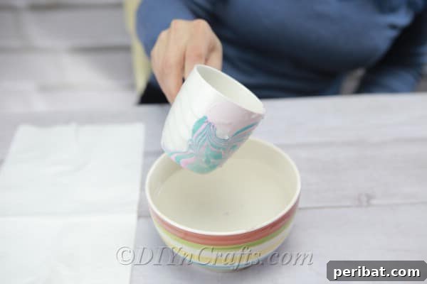 A white coffee cup being carefully dipped into the bowl of water with the marbled nail polish design.