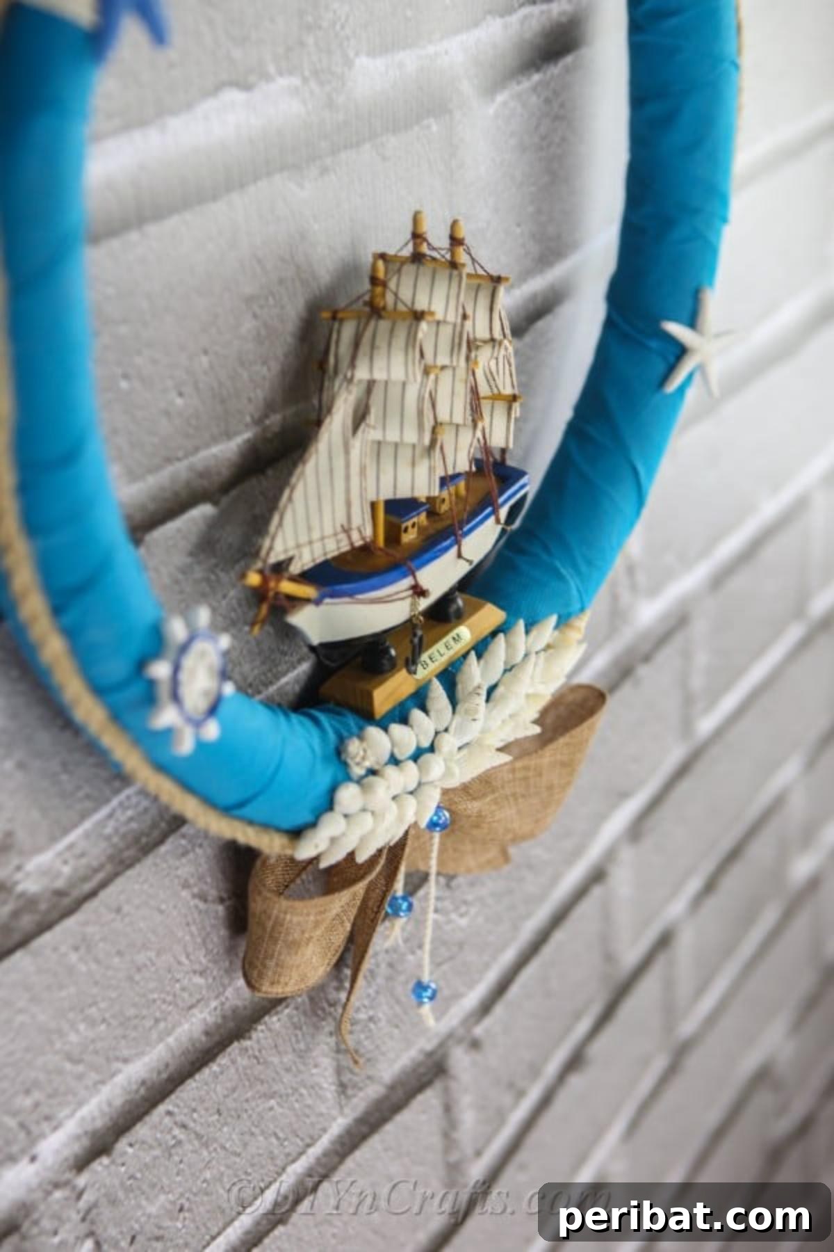 Coastal Elegance: Craft Your Perfect Summer Wreath 5 A close-up view of a beautifully finished DIY nautical wreath, showcasing intricate details like small seashells, a tiny sailboat, and rustic rope, all set against a blue fabric base, hanging on a textured wall.