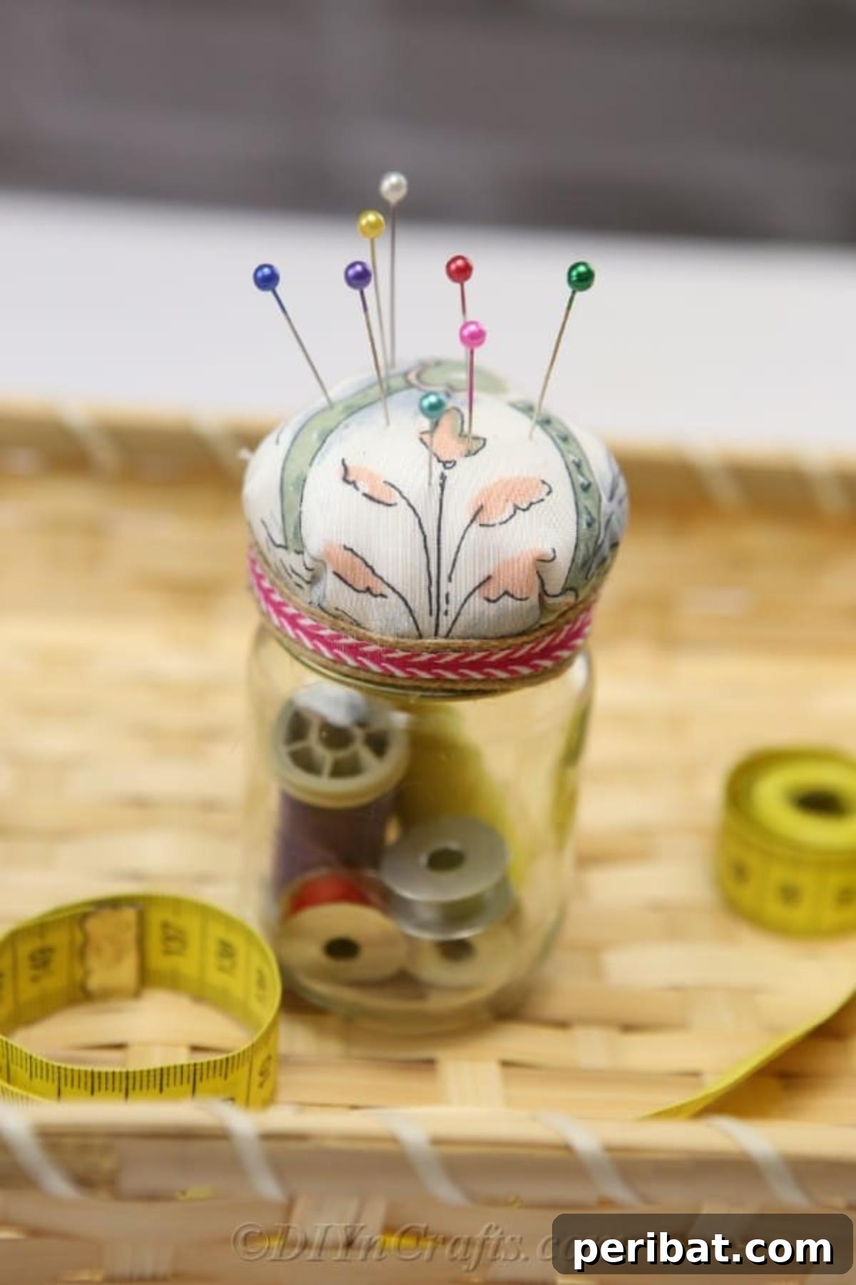 Clever Pincushion Jar Sewing Caddy 22 A completed pincushion jar, featuring a blue floral fabric top and a decorative ribbon, sits among other sewing tools in a wicker basket, ready to be gifted.