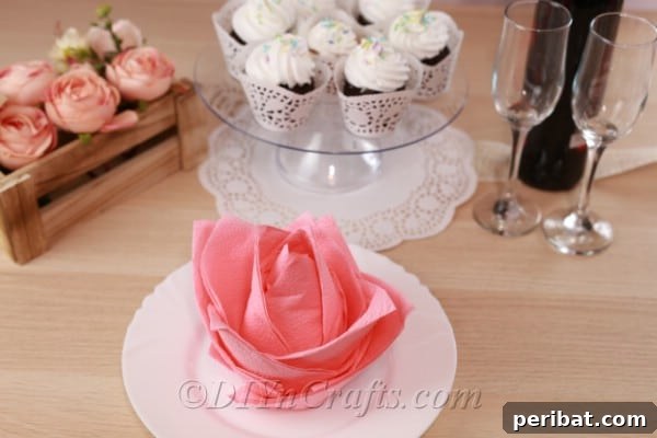 Picture of a flower napkin on a white plate in front of a cake stand with cupcakes and a bottle of wine