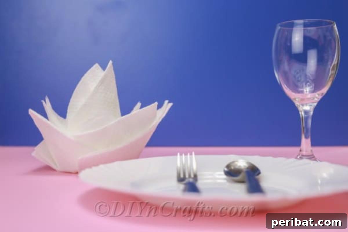 Fold Beautiful Napkin Roses 4 White napkin rose on a pink tablecloth behind a white plate, demonstrating a completed folded napkin