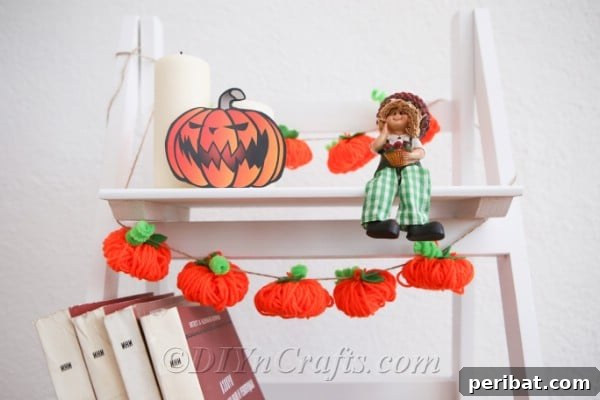 Yarn pumpkin garland diy fall decor hanging on a white shelf next to pumpkins and toy scarecrows