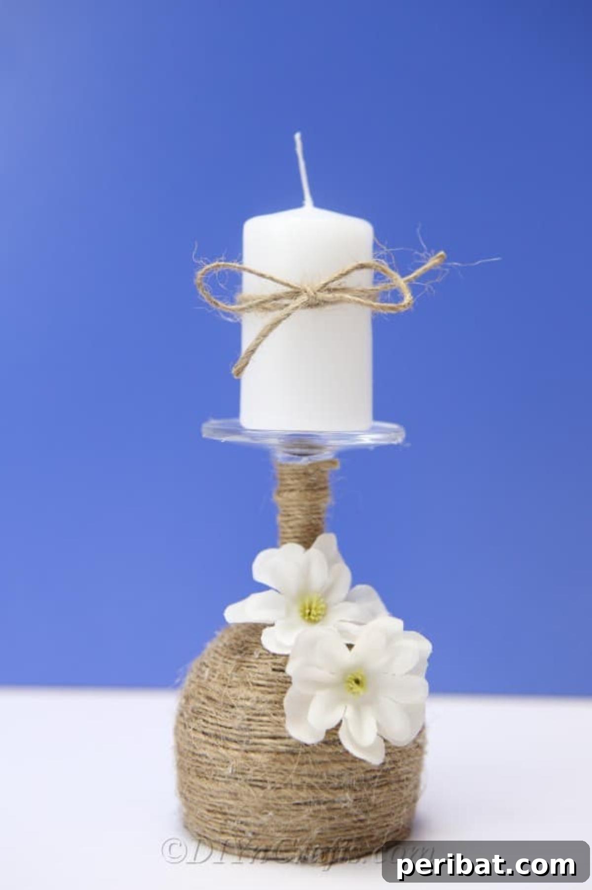 Rustic Glow: DIY Wine Glass Candle Holder 3 A close-up shot of the rustic wine glass candle holder, adorned with twine and faux flowers, casting a gentle glow against a soft blue background.