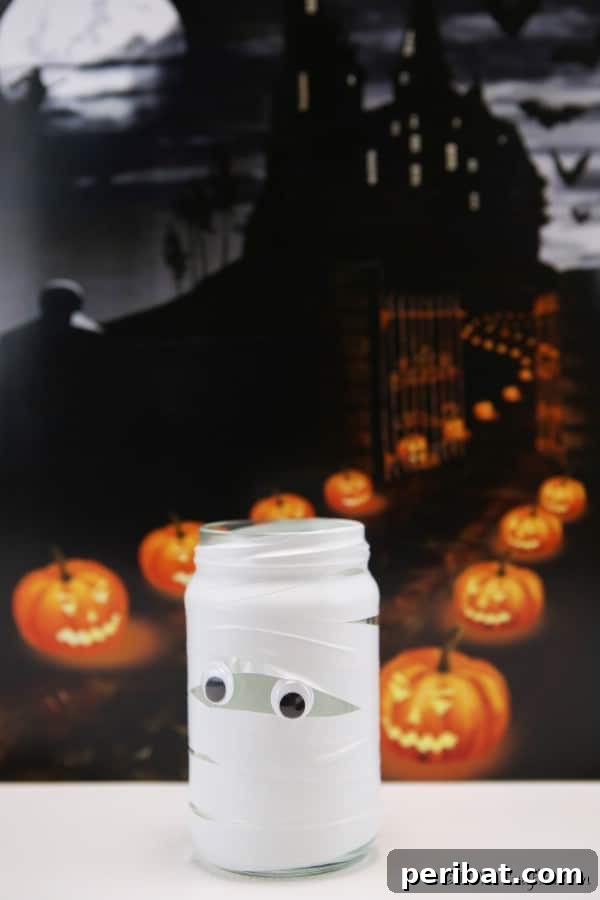 A mummy mason jar light placed outdoors on a white table, with blurred Halloween pumpkins in the background.