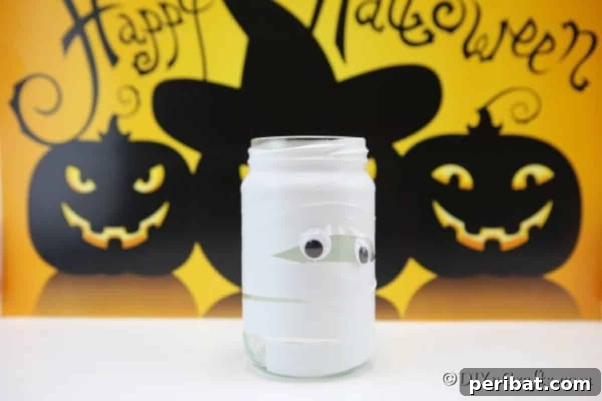 A completed mummy mason jar light with glowing eyes, sitting on a white counter with a festive yellow 'Happy Halloween' banner in the background.