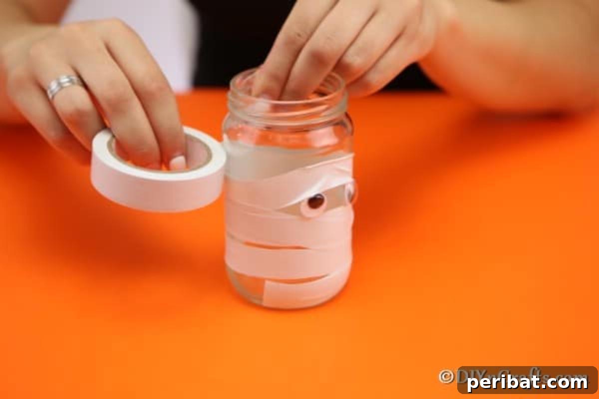 Hands carefully wrapping strips of paper tape around a mason jar, creating the mummy bandage effect, with gaps left strategically.