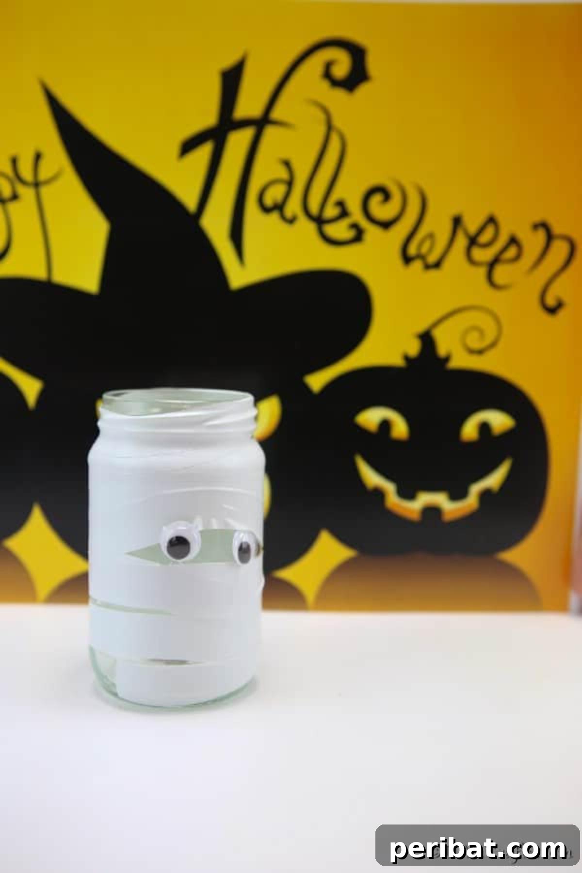 Another perspective of a completed mummy mason jar light, glowing softly on a white counter against a blurred festive Halloween sign in the background.