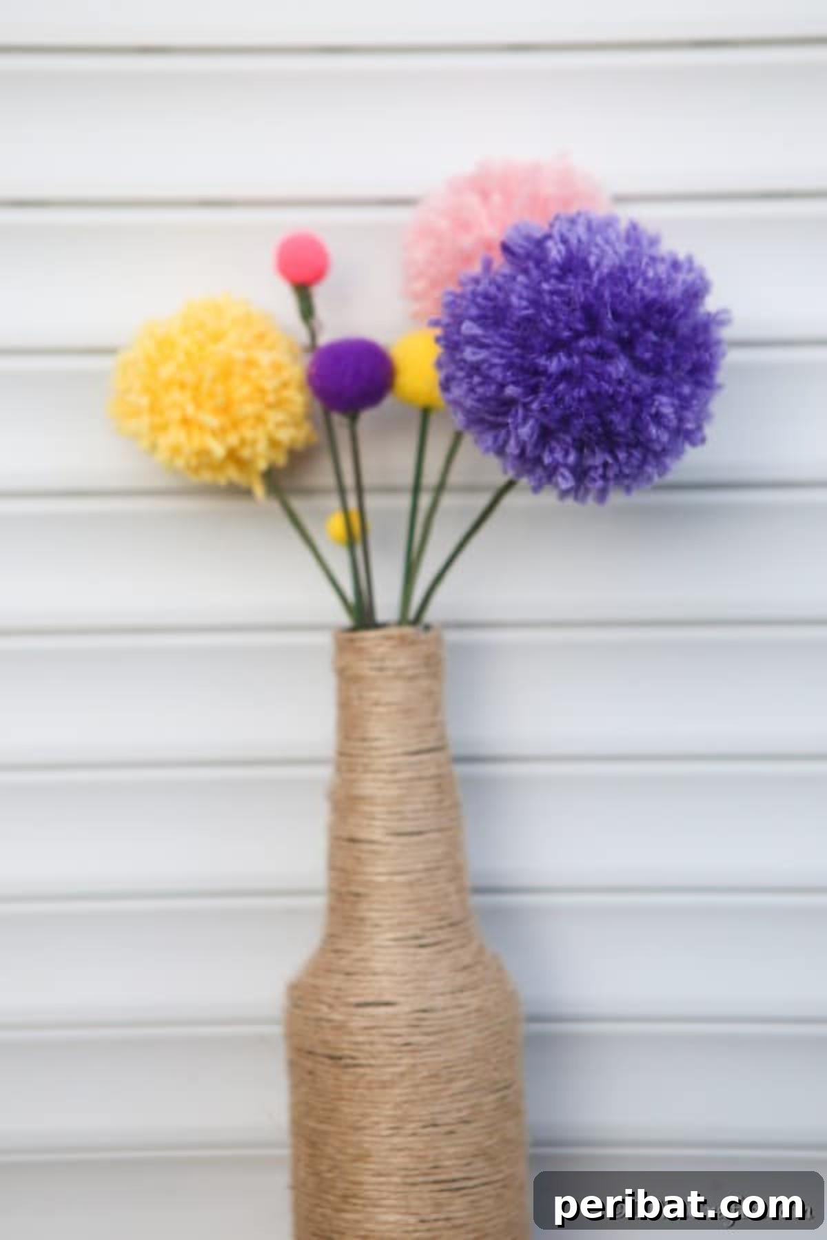 A stylish twine-wrapped vase holds an exquisite arrangement of colorful yarn pom pom flowers, showcasing a blend of red, orange, yellow, and blue hues.