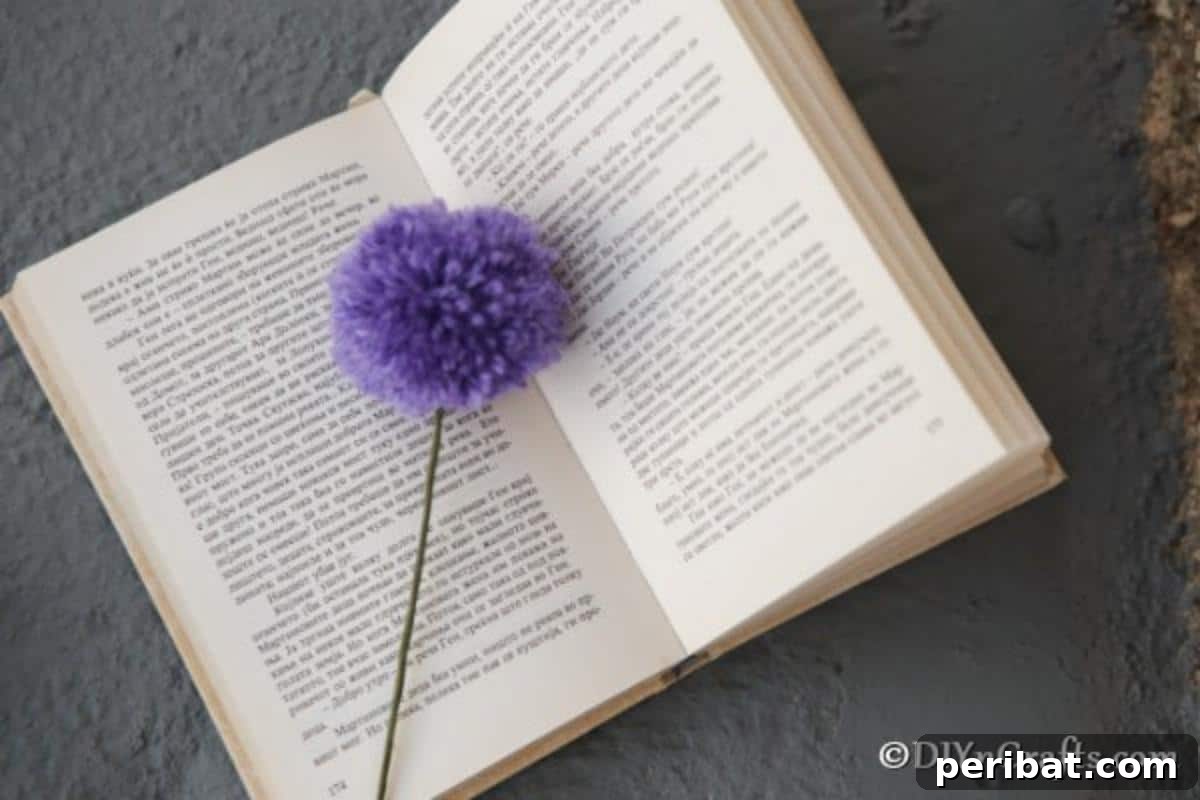 A vibrant purple yarn pom pom flower sits gracefully on the open pages of a book, serving as a charming and functional bookmark.