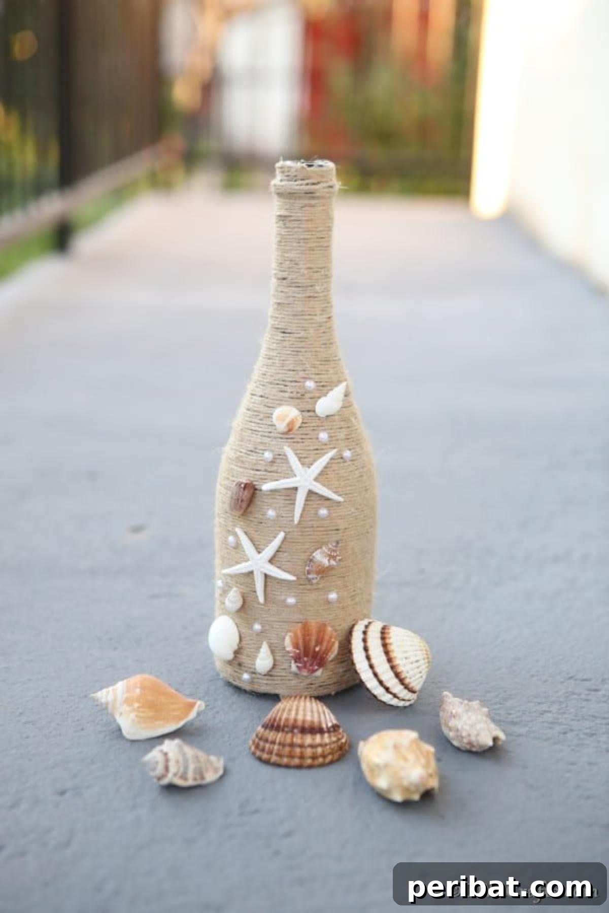 Coastal Rustic Wine Bottle Charm 10 The completed wine bottle decor piece with a rustic nautical theme, adorned with twine and seashells, placed on a concrete walkway.