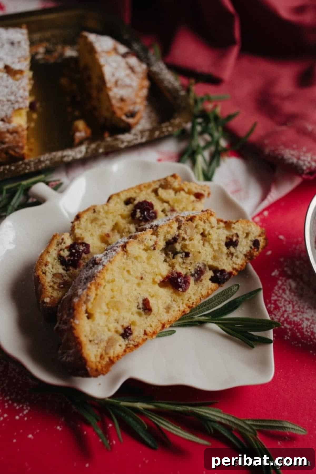 German Christmas Stollen Festive Bread Recipe 6 A beautifully sliced loaf of holiday bread on a white leaf-shaped plate, set against a festive red tablecloth