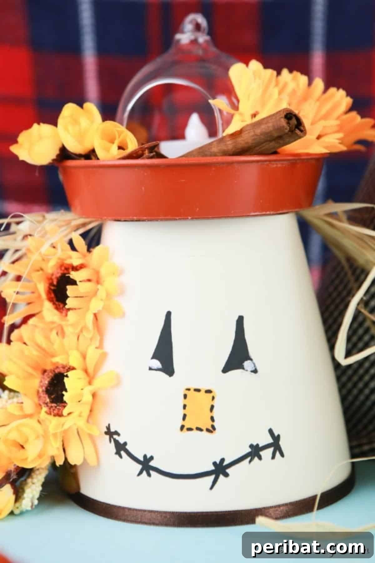 Whimsical Scarecrow Pot Lantern 4 A finished DIY scarecrow clay pot lantern with a whimsical hand-drawn face, ribbon scarf, and hay 'hair', placed on a textured blue surface with a cozy plaid fabric in the background, showcasing its rustic charm.