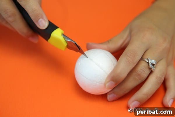 A craft knife carefully scoring the drawn lines on a Styrofoam ball, creating shallow grooves for attaching fabric to make a pumpkin ornament
