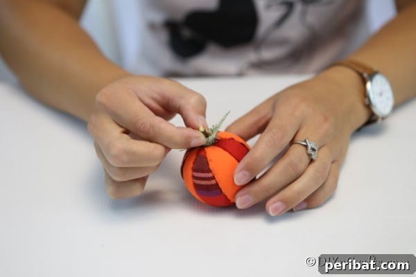 Carefully placing a natural twig into the top center of the fabric pumpkin ornament to create a rustic stem, enhancing its fall appeal