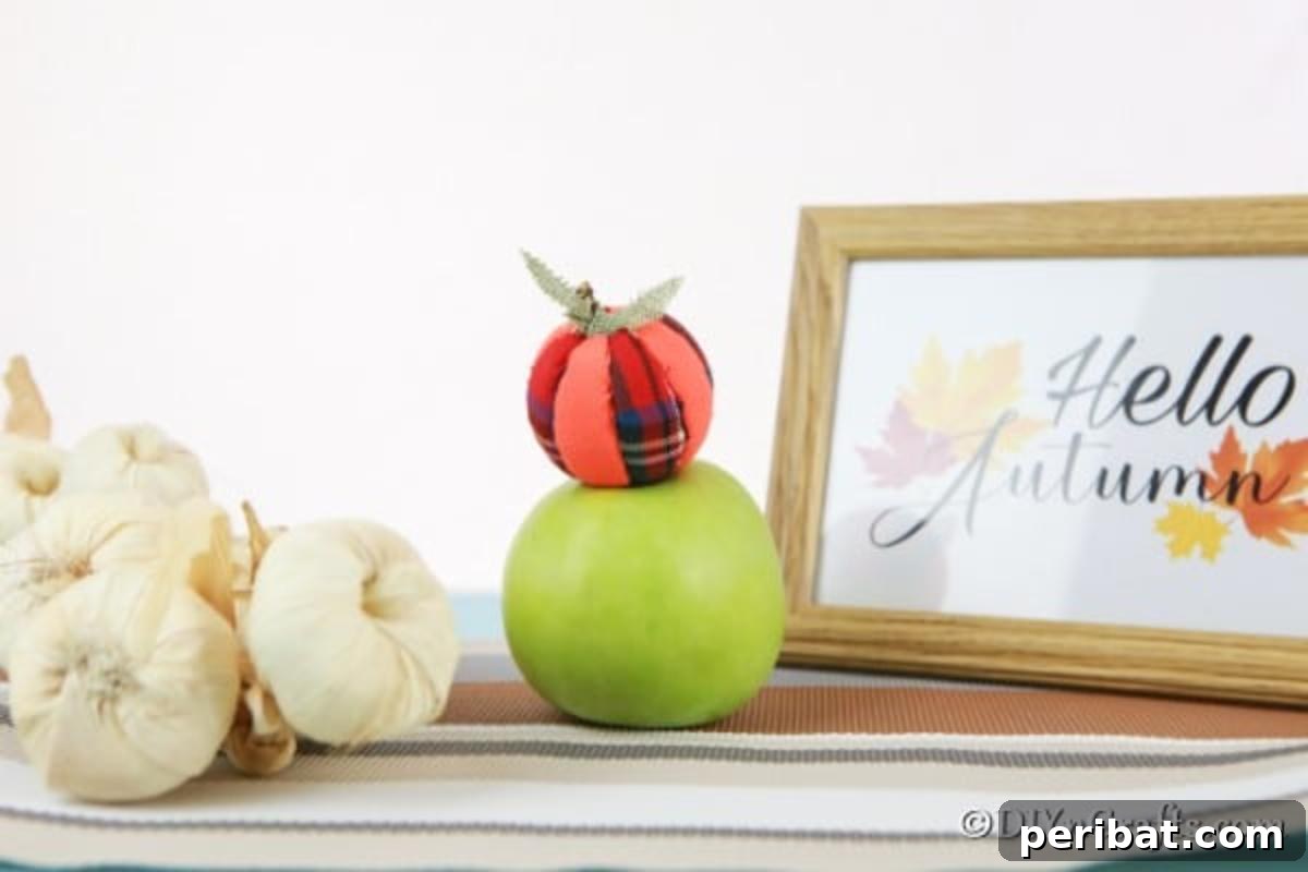 A close-up of a handmade cloth pumpkin ornament resting on a vibrant green apple, with a blurred background featuring a picture and string of garlic, highlighting natural fall elements