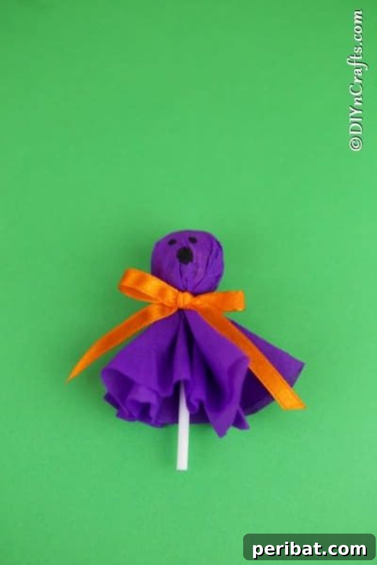 A purple ghost lollipop with a drawn face, resting on a vibrant green surface, ready for Halloween festivities.