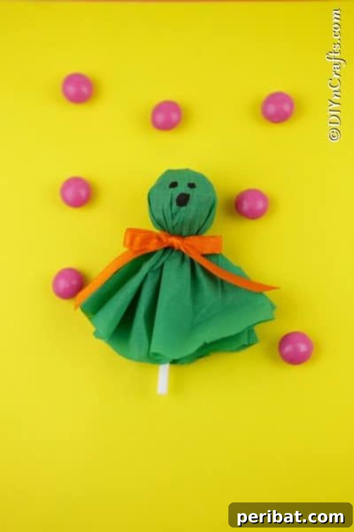 A green ghost craft lollipop with a cheerful face, surrounded by pink gumballs on a yellow surface, radiating Halloween delight.