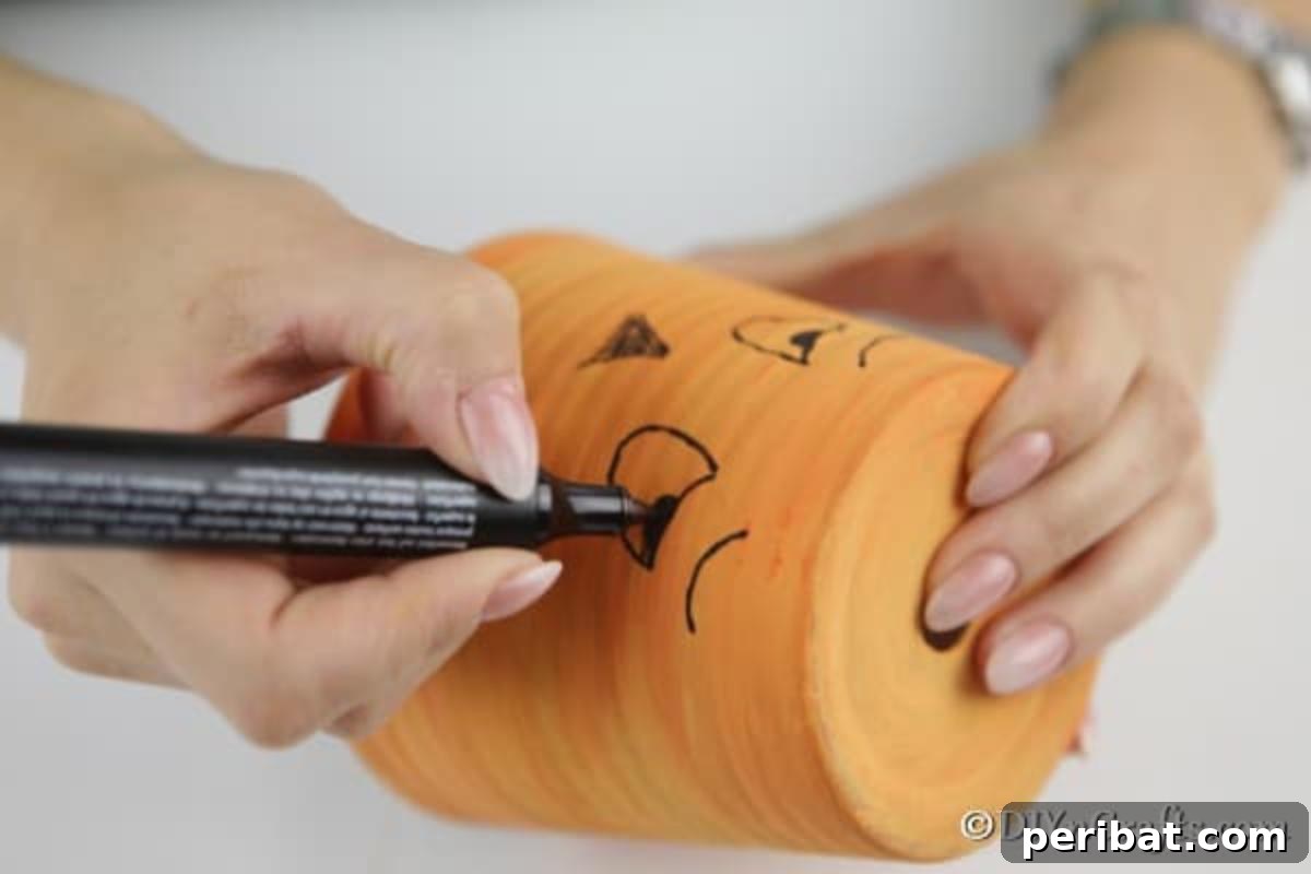 A black paint pen being used to draw the outline of a silly face onto an orange-painted terra cotta pumpkin planter