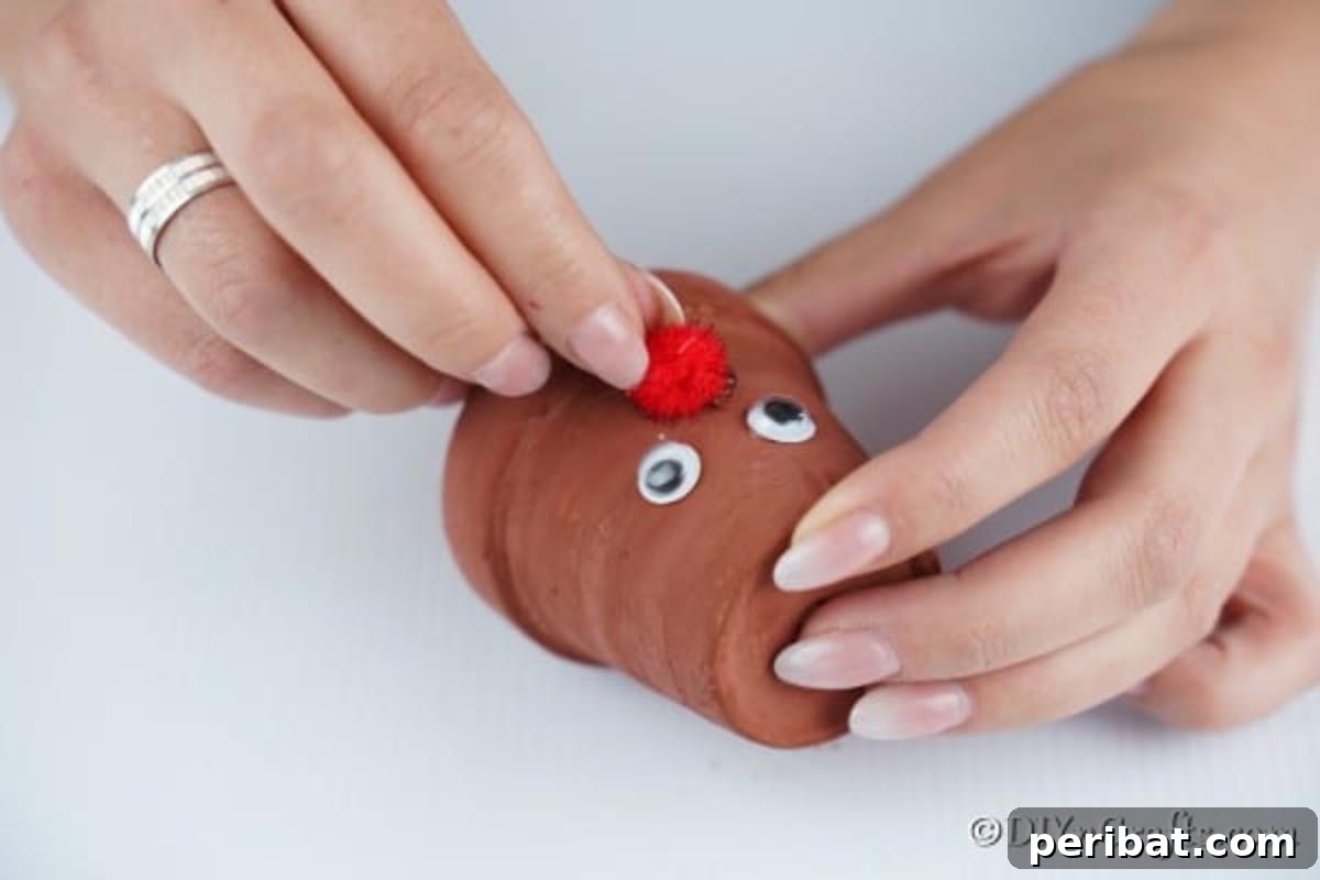 Whimsical Clay Pot Reindeer Christmas Creations 8 Close-up shot of hands carefully adding googly eyes and a bright red pom-pom nose to an inverted small painted flower pot, initiating the creation of a whimsical reindeer Christmas decoration.