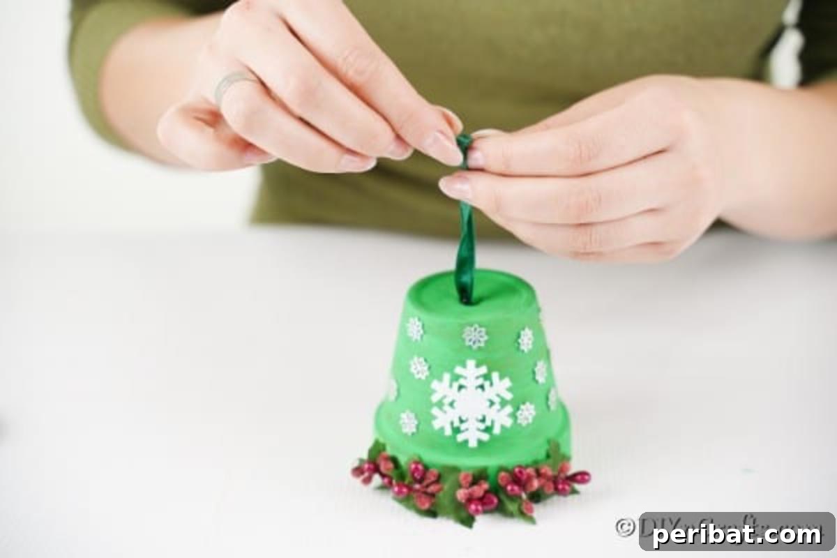 Craft Your Own Adorable Christmas Bell Ornaments 12 A hand pulling a red ribbon with attached jingle bells through the top hole of a green and decorated clay pot, completing the assembly of the Christmas bell decoration