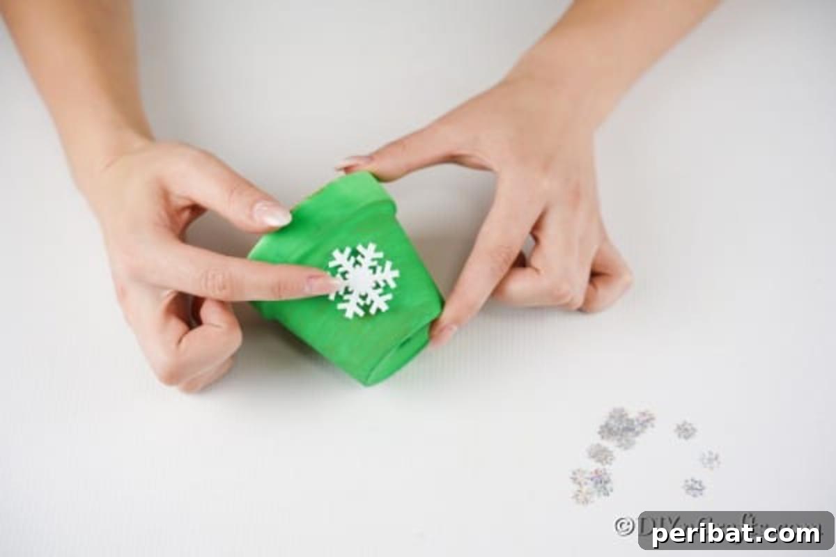 Craft Your Own Adorable Christmas Bell Ornaments 8 Close-up of a hand carefully gluing a white snowflake embellishment onto the green painted surface of a clay pot, transforming it into a Christmas bell