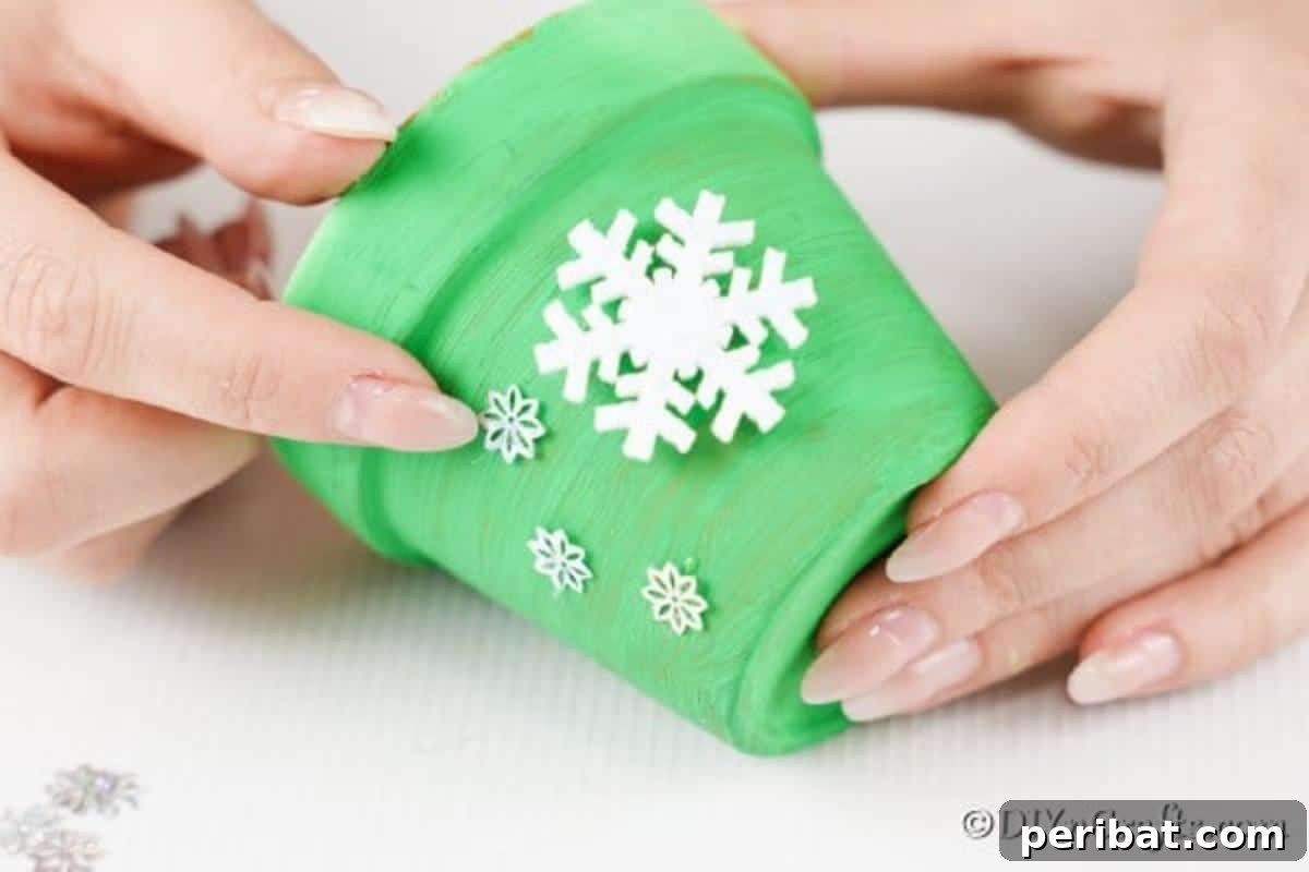Craft Your Own Adorable Christmas Bell Ornaments 9 A hand applying multiple white snowflake decorations to the side of a green painted clay pot, enhancing its appearance as a holiday bell craft