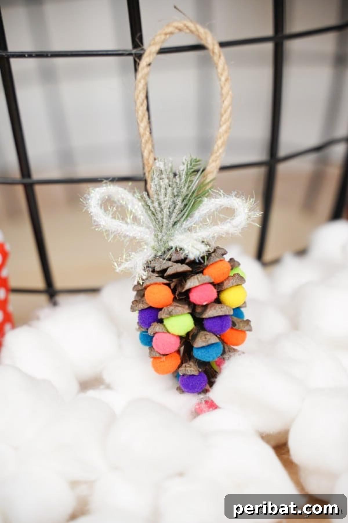 Vibrant Pinecone Christmas Decorations 5 A single colorful pinecone ornament with vibrant pom poms hanging delicately above a bed of fluffy cotton balls, creating a cozy winter scene.