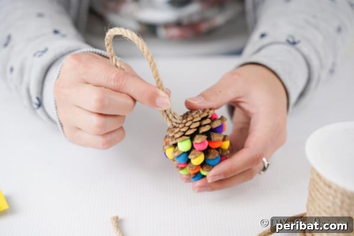 Vibrant Pinecone Christmas Decorations 8 A hand adding a loop of natural twine, secured with glue, to the top of a pom-pom adorned pinecone to create the ornament hanger.