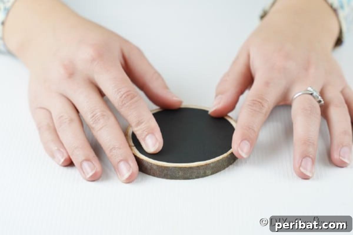 Rustic Wood Slice Chalkboard Charm 9 Close-up of a hand carefully applying a round piece of black adhesive chalkboard paper onto the flat surface of a rustic wood slice, ensuring a smooth and centered application for the ornament base.