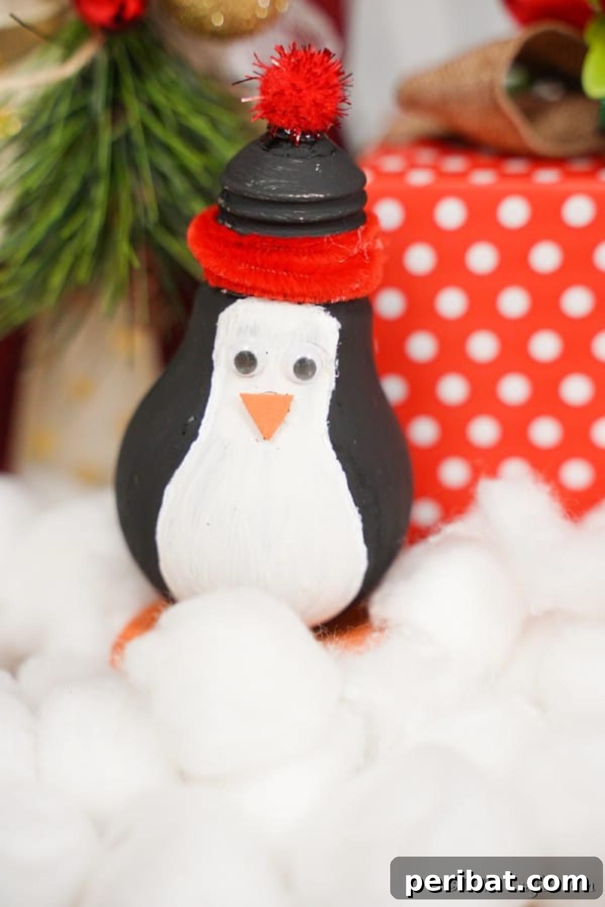 A light bulb penguin sitting in front of a red and white present and on top of cotton balls