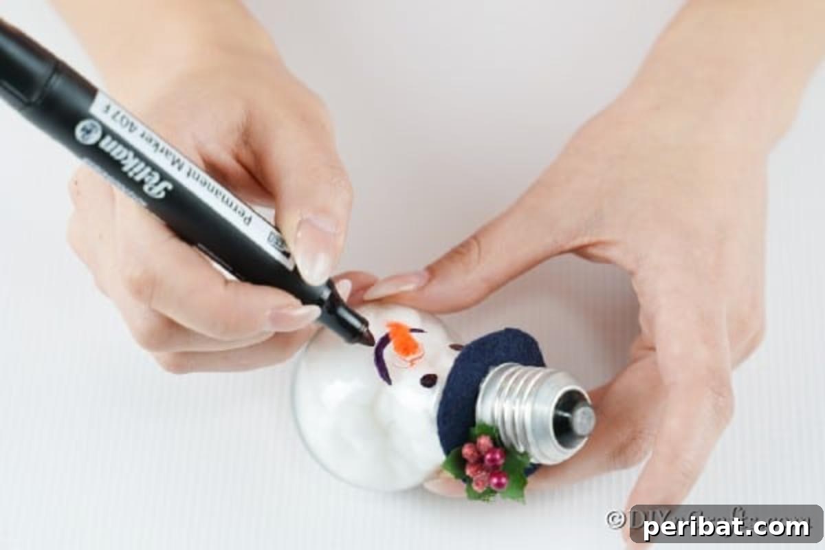 A close-up shot of the light bulb snowman ornament as its face is being completed, showing the attached orange fabric nose and drawn-on eyes and mouth
