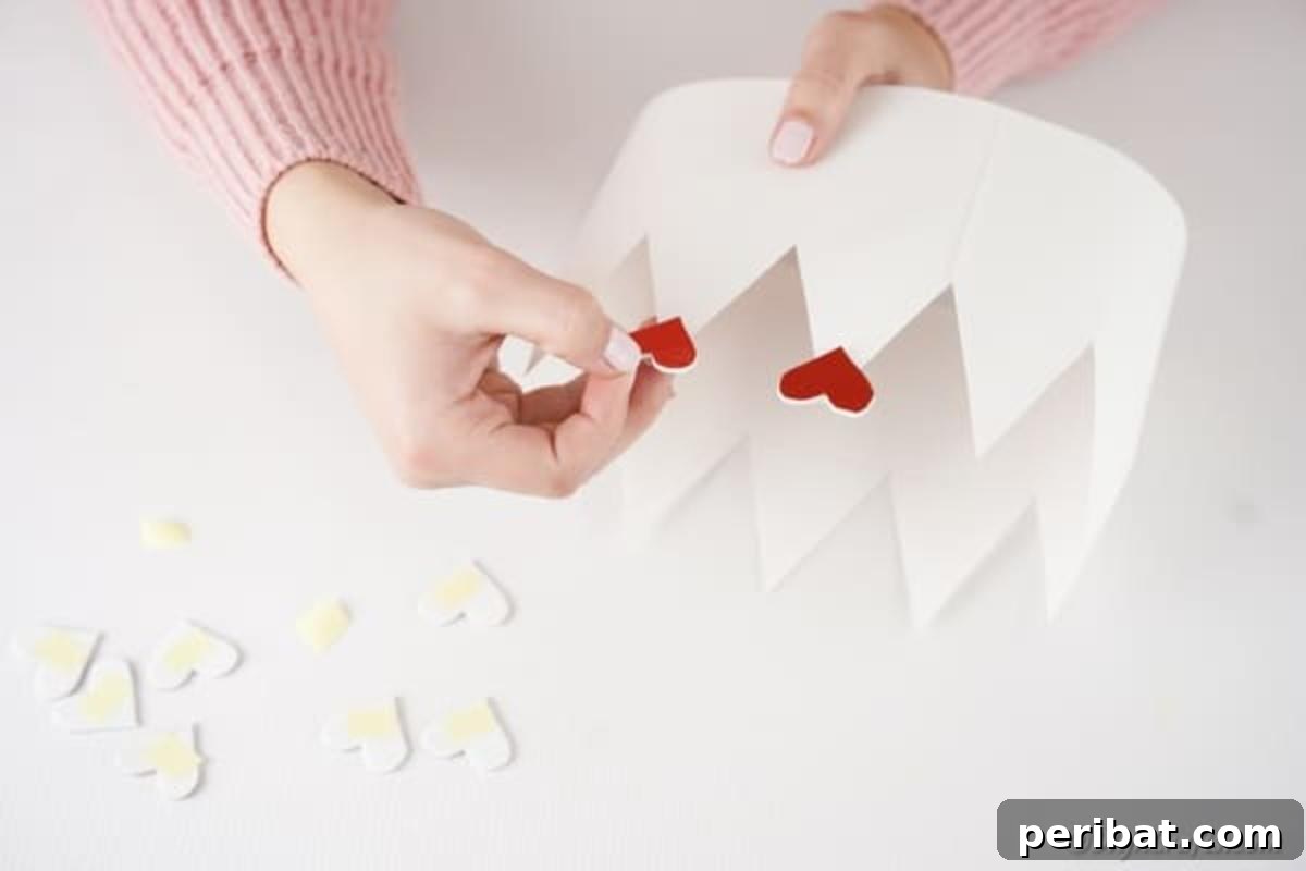 Easy Valentine Paper Crown Craft for Children 11 A hand glues a large heart decoration to the top of a crown peak, adding a primary embellishment to the paper crown.